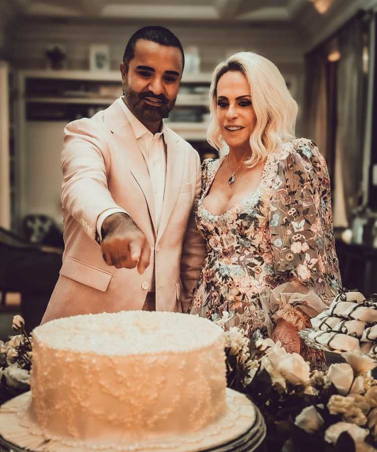 Casal Ana Maria Braga e Fabio Arruda elegante sorri enquanto corta bolo decorado com flores brancas em celebração íntima. Noiva veste vestido floral romântico em tons pastel e noivo usa terno claro. Cenário acolhedor com iluminação quente e decoração requintada complementa o momento especial.
