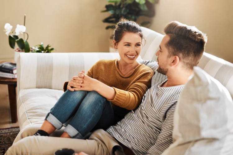 Casal heterossexual, de pele clara, sentado no sofá conversando e sorrindo