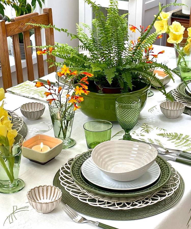 Mesa posta com decoração vibrante: samambaia central em vaso verde cercada por flores amarelas e laranjas. Aparelho de jantar com louça branca e verde, copos de vidro, guardanapos com estampa de folhas e vela acesa criam um ambiente elegante e acolhedor.