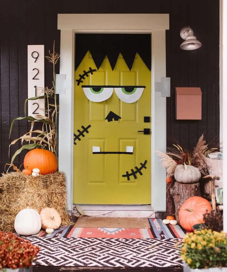 Decoração de entrada com parede marrom, porta verde com desenho de Frankenstein, feno e abóboras