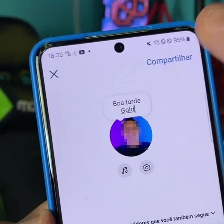 A imagem mostra a tela de um smartphone exibindo um perfil no Instagram. No centro da tela, há uma mensagem de saudação que diz "Boa tarde Gold". Abaixo da mensagem, há a foto de perfil do usuário, que está pixelada para preservar a privacidade. Ao redor da foto de perfil, há ícones de música e câmera. No canto superior direito, há a opção "Compartilhar". A mão de uma pessoa segura o telefone, que está em uma capa azul. Ao fundo, é possível ver um ambiente desfocado com uma cadeira e um objeto decorativo.


