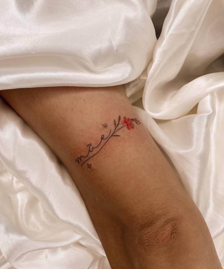 Uma delicada tatuagem da palavra "mãe" com um ramo de flores vermelhas e pequenos brilhos está no antebraço de uma pessoa. O braço repousa sobre um tecido branco amassado, realçando o design minimalista da tatuagem.