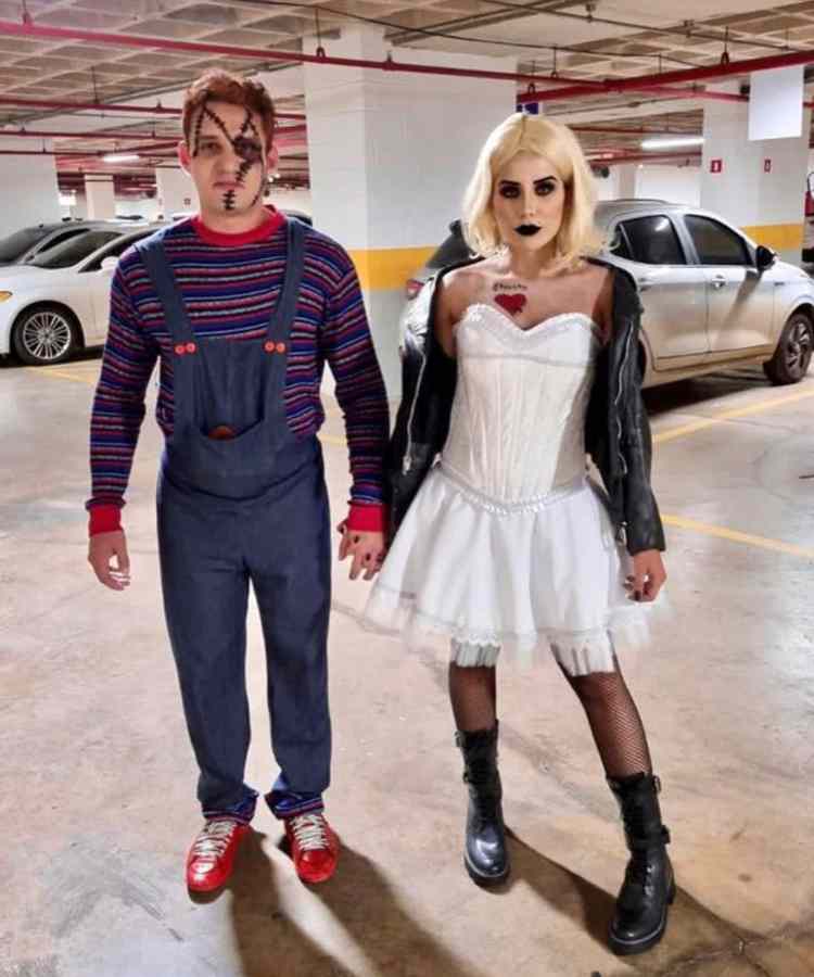 Fantasia de casal para o Halloween: Chucky e Tiffany. Ele usa blusa colorida, jardineira longa e cicatrizes no rosto. Ela usa cabelo loiro curto, jaqueta preta, vestido branco e batom escuro 