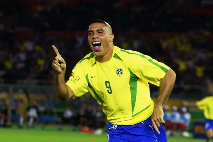 A imagem mostra um jogador de futebol vibrando intensamente, vestindo o uniforme da seleção brasileira, com camisa amarela, shorts azuis e número 9. Ele aponta para o alto, sorrindo, com fundo desfocado de estádio e torcida ao fundo.
