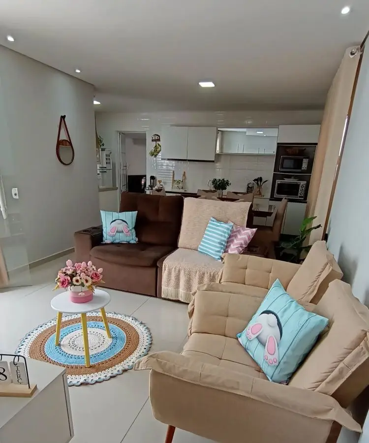 Sala de estar integrada com cozinha, sofás bege e marrom decorados com almofadas temáticas de coelho. Mesa de centro com arranjo de flores, tapete de crochê colorido, ambiente iluminado e moderno com detalhes aconchegantes e organizados.