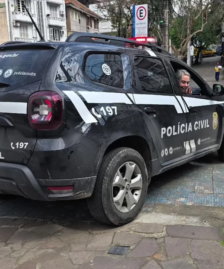 Carro da Polícia Civil do Rio Grande do Sul estacionado em via urbana, com sirene ligada. Veículo preto e branco, identificado pelo número 197, exibe adesivos institucionais. No banco do motorista, uma pessoa aparece sorrindo e acenando.