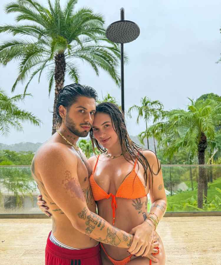 A imagem mostra um casal abraçado em um ambiente tropical, cercado por vegetação com palmeiras ao fundo. Ambos estão molhados, sugerindo chuva ou piscina. A mulher usa biquíni laranja vibrante, e o homem exibe tatuagens no corpo.


