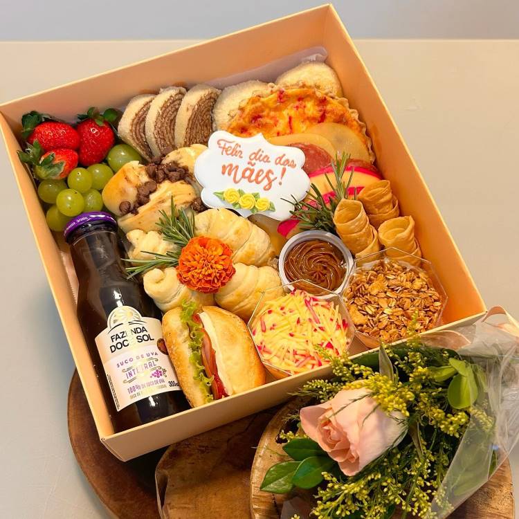 Cesta com frutas, pães, salgados, doces, granola e doce de leite. Decoração inclui flor e mensagem "Feliz Dia das Mães". Acompanha suco de uva em garrafa e um pequeno buquê com rosa. Apresentação elegante e acolhedora.