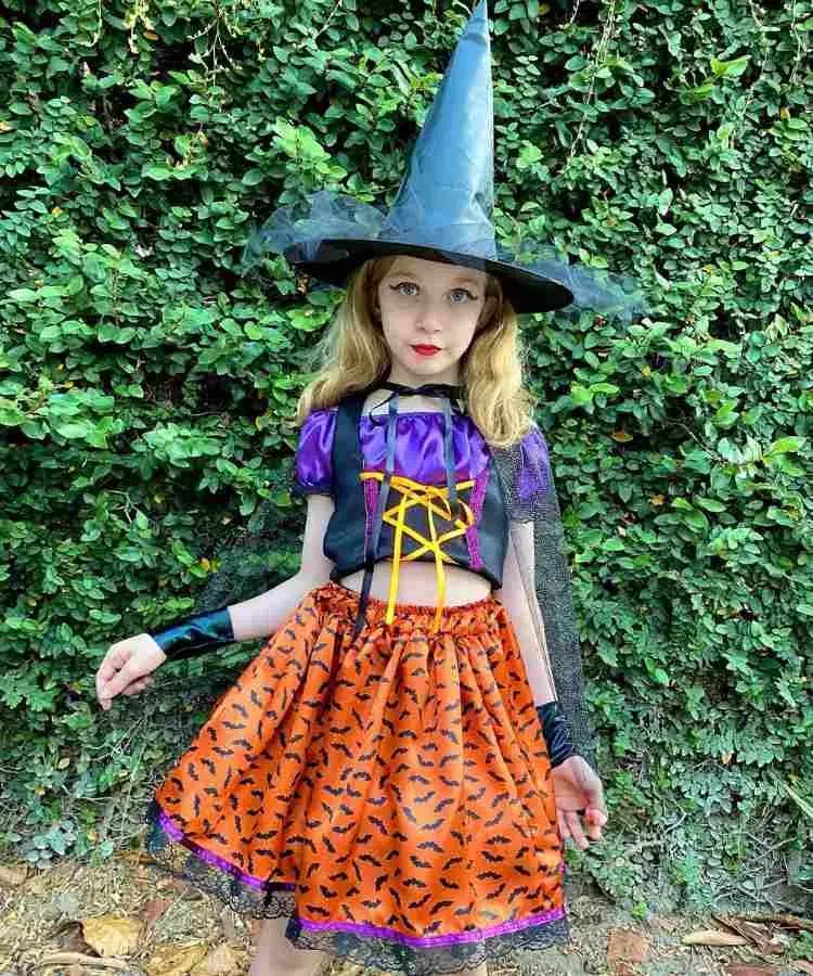 Garota com fantasia de bruxa infantil. Cores predominantes: roxo, laranja e preto. Usa: croped, saia, luvar, chapéu pontudo e capa