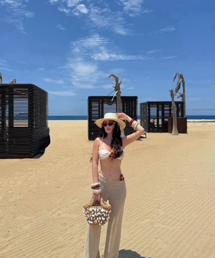 Mulher em pé em uma praia de areia, usando uma saída de praia de crochê bege, chapéu de palha e óculos de sol. Ela segura uma bolsa e posa com a mão na cabeça.