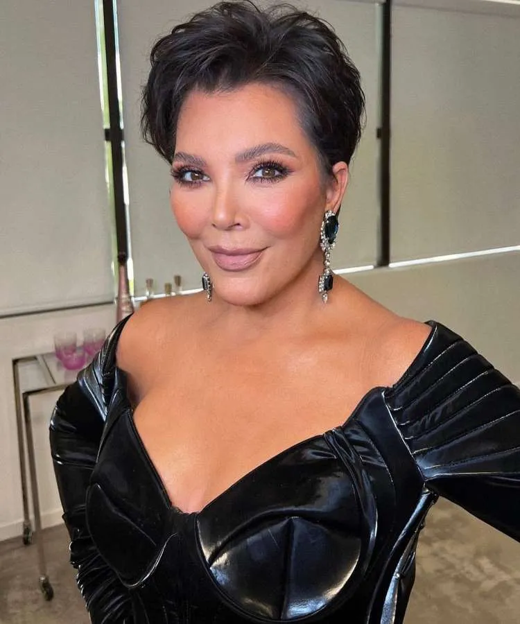 Kris Jenner sorri em um bustier preto brilhante, com brincos grandes e escuros. Seu cabelo curto e escuro é estilizado com volume, e sua maquiagem inclui um batom nude.