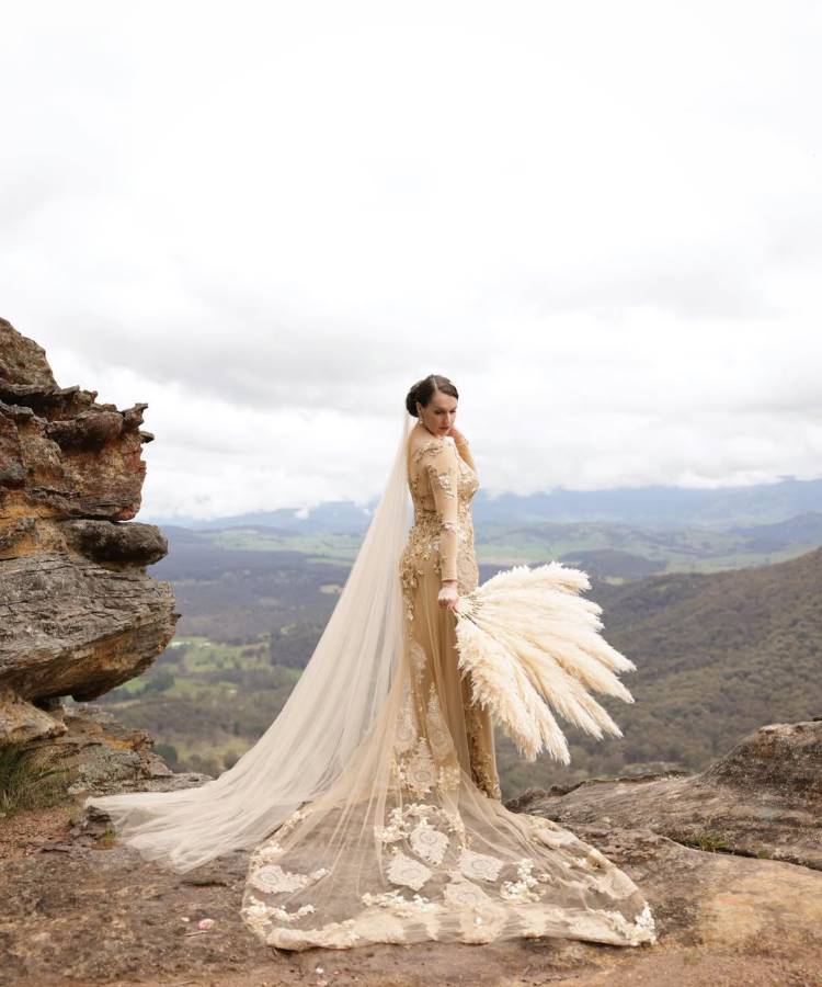 Uma noiva com coque baixo, usando um deslumbrante vestido de noiva dourado e rendado de mangas compridas, com véu longo e um buquê de plumas claras, posa em um penhasco rochoso com montanhas e céu nublado ao fundo.







