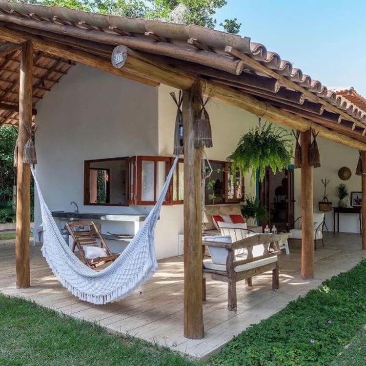 Imagem de entrada de lar, com alpendre, que mostra uma casa branca com bancada de pia, brancos de madeira, rede e planta samambaia pendurada