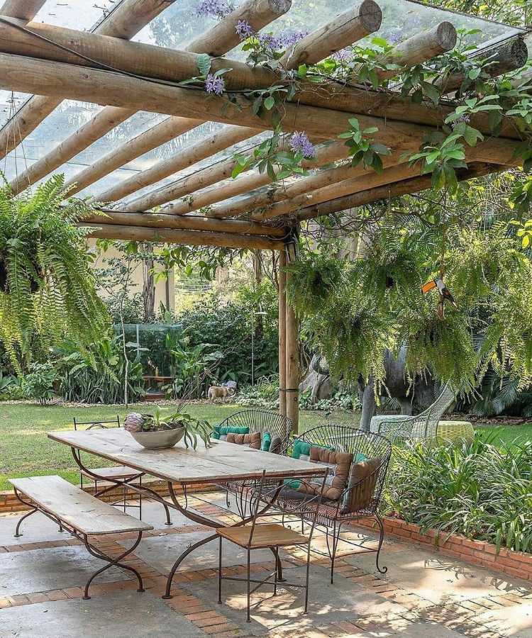 Pergolado em área externa com diversas plantas, incluindo samambaia, com mesa de madeira e ferro abaixo