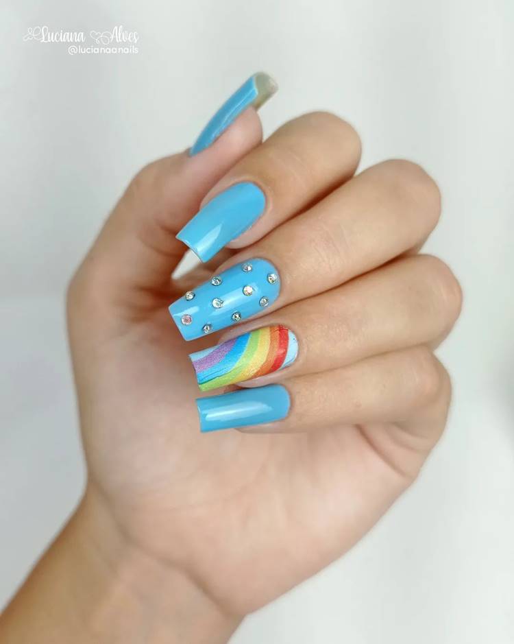 Mão com unhas arco-íris LGBTQIA+: Polegar: azul céu Indicador: azul céu Médio: azul céu com pedrinhas Anelar: azul céu com ar-íris e glitter Mínimo azul céu