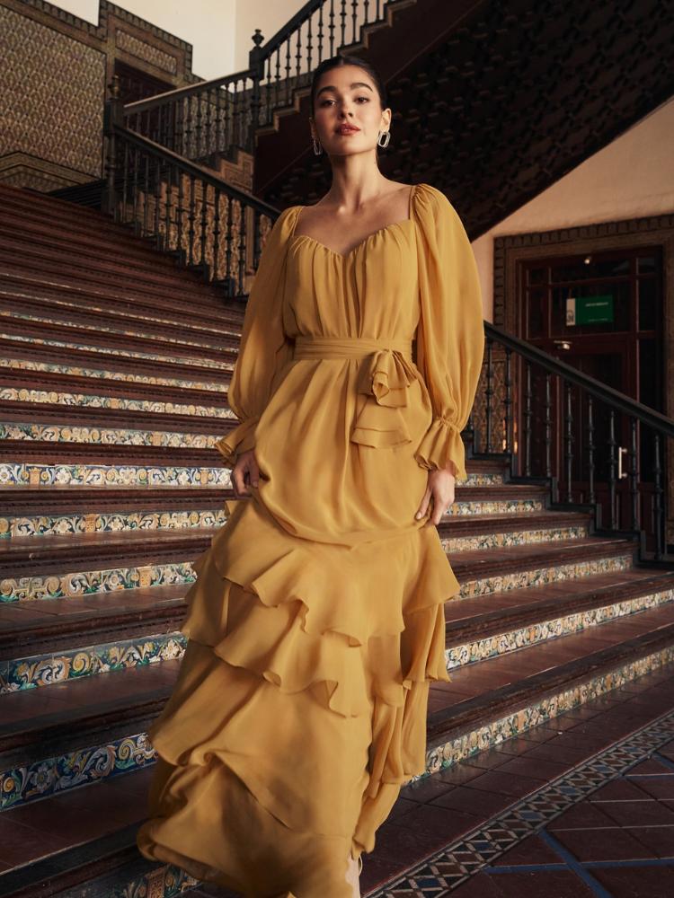 Mulher de pele clara usando vestido mostarda com babados e laço discreto na cintura 