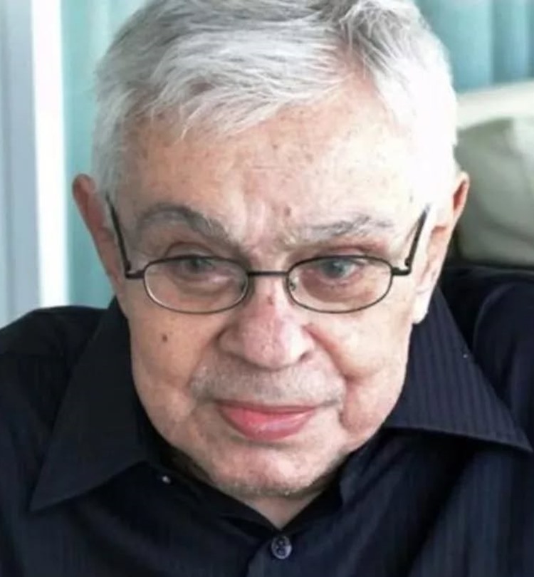 Close-up de um homem branco de idade com cabelos grisalhos e óculos. Ele usa uma camisa de botão escura e olha diretamente para a câmera com uma expressão séria. O fundo está desfocado.