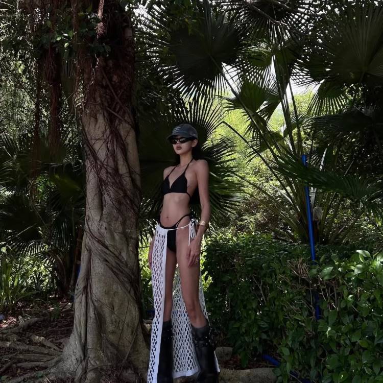 Mulher magra posa em pé perto de uma árvore e vegetação. Ela usa um biquíni preto, chapéu, óculos de sol, botas e uma saída de praia de crochê branca transparente sobre o biquíni.