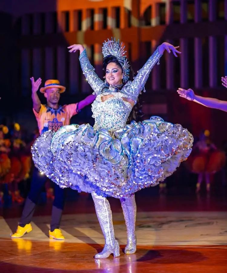 Imagem de uma rainha da quadrilha em festa junina, em um palco, com um vestido brilhante prateado e azul, saia rodada e botas. Ela sorri enquanto dança com os braços erguidos, cercada por outros dançarinos ao fundo. Fontes
