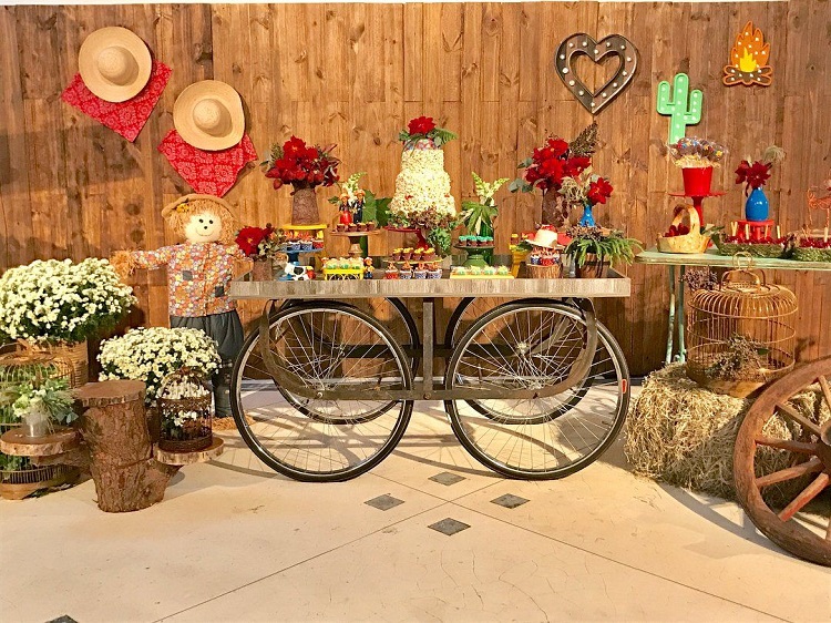 A imagem representa decoração para festa junina com mesas diferentes, usando rodas de bicicleta. Contém doces, bandeirinhas, arranjos de flores, espantalhos, feno, gaiola, chapéus de palha, etc