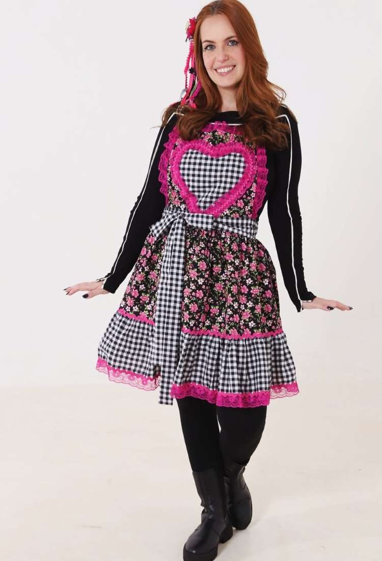 Uma mulher ruiva sorri vestindo um avental junino sobre roupa preta. O avental possui um grande coração xadrez rosa e preto no busto, e a saia florida preta com detalhes xadrez e renda rosa. Botas pretas e um laço vermelho no cabelo completam o visual.


Fontes






