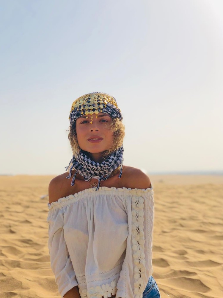 Larissa usando blusa branca ombro a ombro, lenço na cabeça, no deserto em Dubai