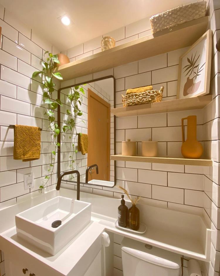 Banheiro branco com luz amarela, plantas, pia branca, espelho, vaso sanitário, decorações em tons terrosos e prateleiras