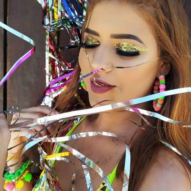 Mulher de pele clara com sombra dourada de glitter, batom rosa, brincos coloridos e em meio a papéis metalizados