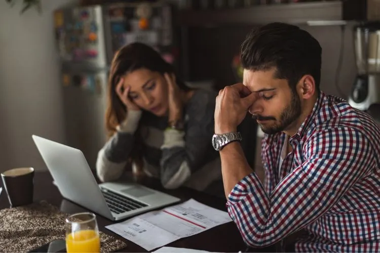 Um casal parece preocupado enquanto olha para papéis e um laptop em uma mesa. O homem, em primeiro plano, esfrega a testa, enquanto a mulher, desfocada ao fundo, apoia a cabeça na mão. A cena sugere a busca por soluções para sair do vermelho financeiramente.

