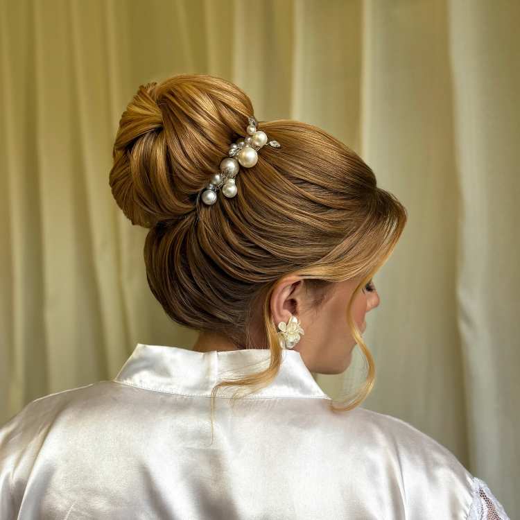 Coque alto com cabelo loiro volumoso, decorado com pérolas e acessórios brilhantes, usada para ocasião formal, com robe de cetim branco.