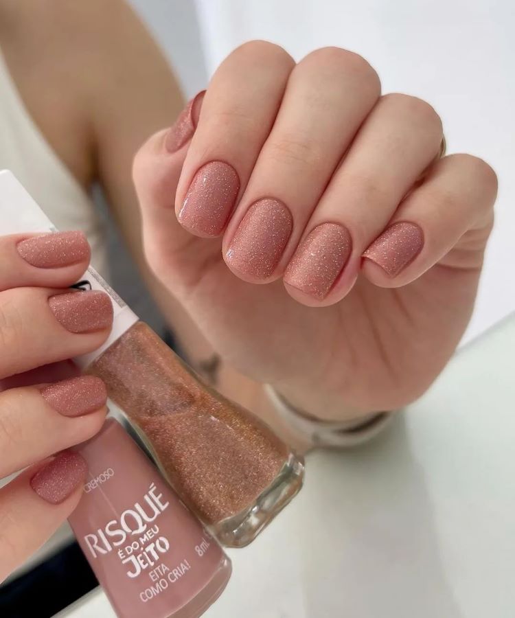 A imagem mostra uma mão com unhas pintadas em um tom de rosa cintilante, todas uniformemente decoradas, criando um visual elegante e discreto. O esmalte utilizado é da marca Risqué, evidenciado pelo frasco segurado na outra mão. Este estilo é um exemplo de unha decorada simples, destacando-se pela sutileza e brilho suave.
