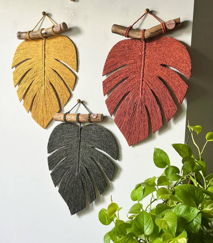 três folhas decorativas de cores diferentes (amarela, vermelha e cinza) penduradas na parede. As folhas são feitas de material têxtil e estão penduradas em hastes de madeira que têm pequenos pedaços circulares de madeira anexados a elas. Ao lado das folhas, no canto inferior direito da imagem, há uma planta com folhas verdes. A parede ao fundo é branca, o que ajuda a destacar as cores vibrantes das folhas decorativas.