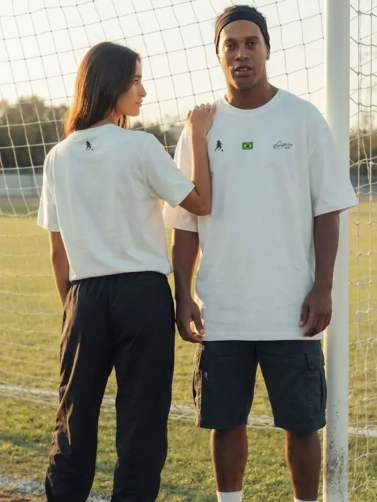 Ronaldinho Gaúcho usando camiseta branca oversized de sua marca de roupas ao lado de modelo