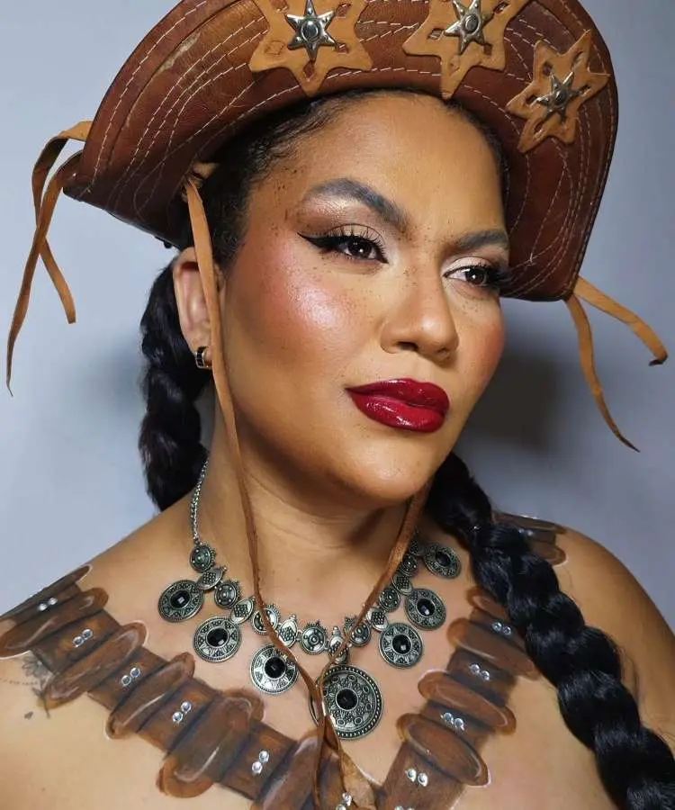 Uma mulher com maquiagem inspirada em Festa Junina, com um chapéu de couro e um delineado marcante. Ela usa um batom vermelho brilhante e possui sardas falsas. Seu corpo parece ter pintura imitando um colete.