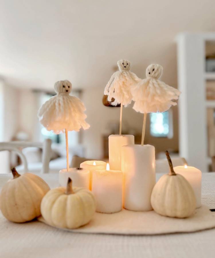 A imagem apresenta uma decoração de Halloween com pequenos fantasmas de lã branca em palitos, posicionados sobre velas brancas de tamanhos variados. Ao redor, há pequenas abóboras brancas. O ambiente é iluminado e acolhedor, com um fundo desfocado, criando uma atmosfera suave e festiva.


