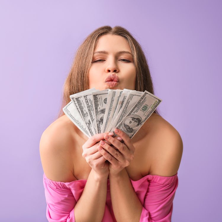 A imagem mostra uma mulher posando contra um fundo roxo, segurando leques de notas de dólar com expressão alegre e confiante. O cenário transmite prosperidade e segurança, destacando a importância de estratégias para alcançar investimentos blindados e financeiramente protegidos.