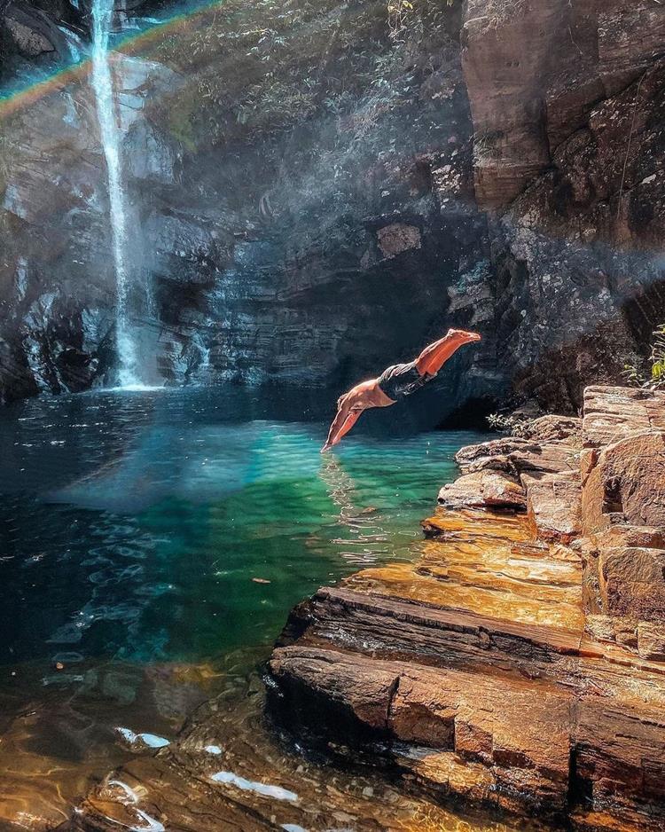 Foto da cidade de cachoeira em Pirenópolis, com homem de pele clara usando bermuda preta pulando na água em uma caverna