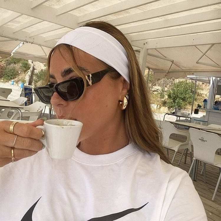 Mulher com cabelo castanho liso solto, faixa branca, óculos escuros e brincos dourados, tomando café ao ar livre em um local arejado.