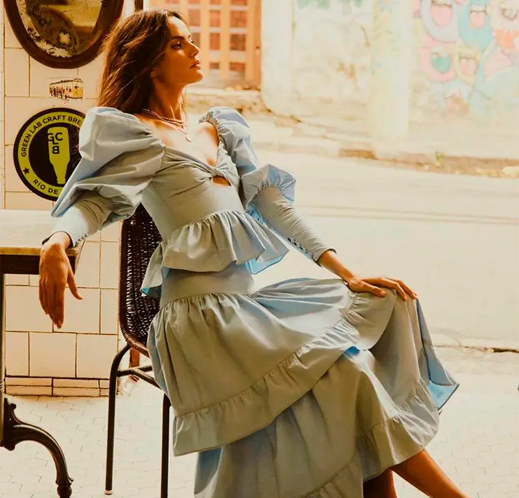 Modelo senta em cadeira e usa um vestido azul claro longo com mangas.