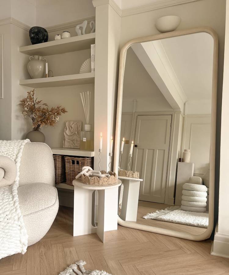 A imagem mostra um ambiente acolhedor em tons neutros, com destaque para um espelho grande de moldura bege encostado na parede. Há uma poltrona confortável com manta de tricô, mesa lateral com cestos e velas, e prateleiras decoradas com cerâmicas e livros. O piso de madeira clara complementa o espaço.