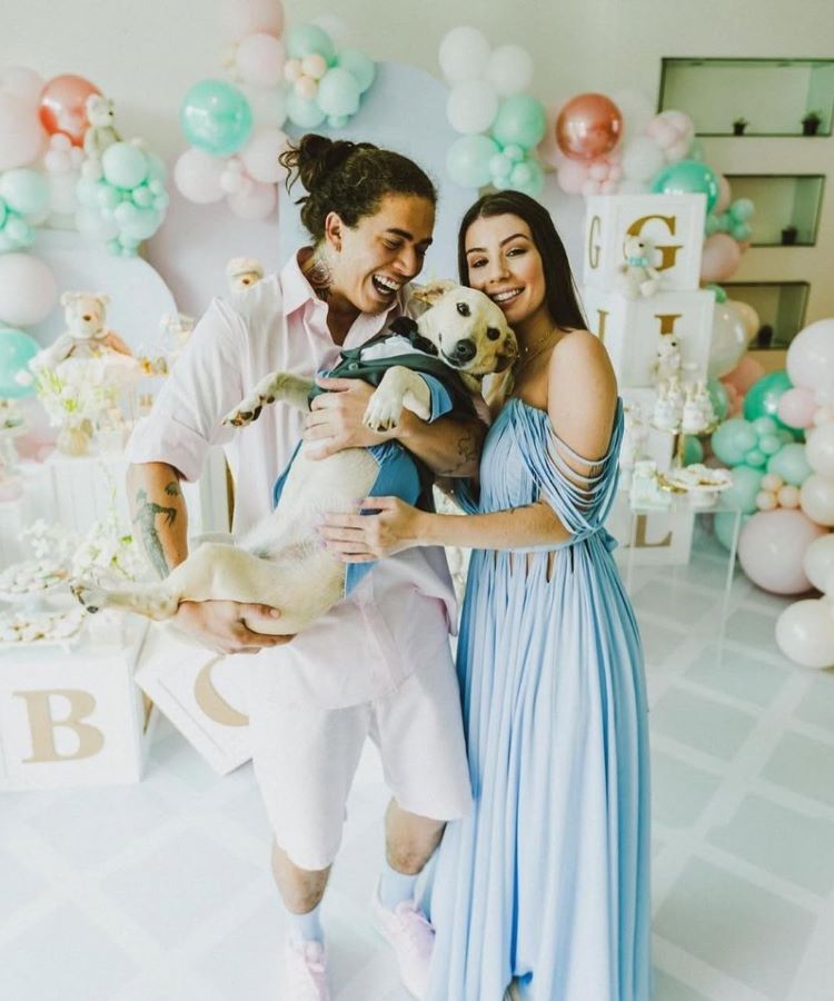 Whindersson Nunes e Maria Lina segura um cachorro vestido com roupa elegante, em um ambiente decorado com balões coloridos em tons pastéis. Ambos estão bem arrumados, ele de camisa rosa e ela em vestido azul longo. Decoração remete a festa infantil.