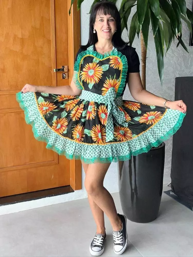 Mulher de pele clara e cabelo escuro usando blusa preta e avental junino com flores amarelas e babado azul