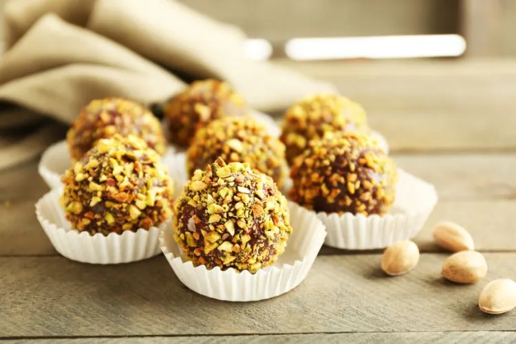 7 Brigadeiros de pistache em forminhas brancas sob superfície de madeira com 4 sementes da oleaginosa ao lado