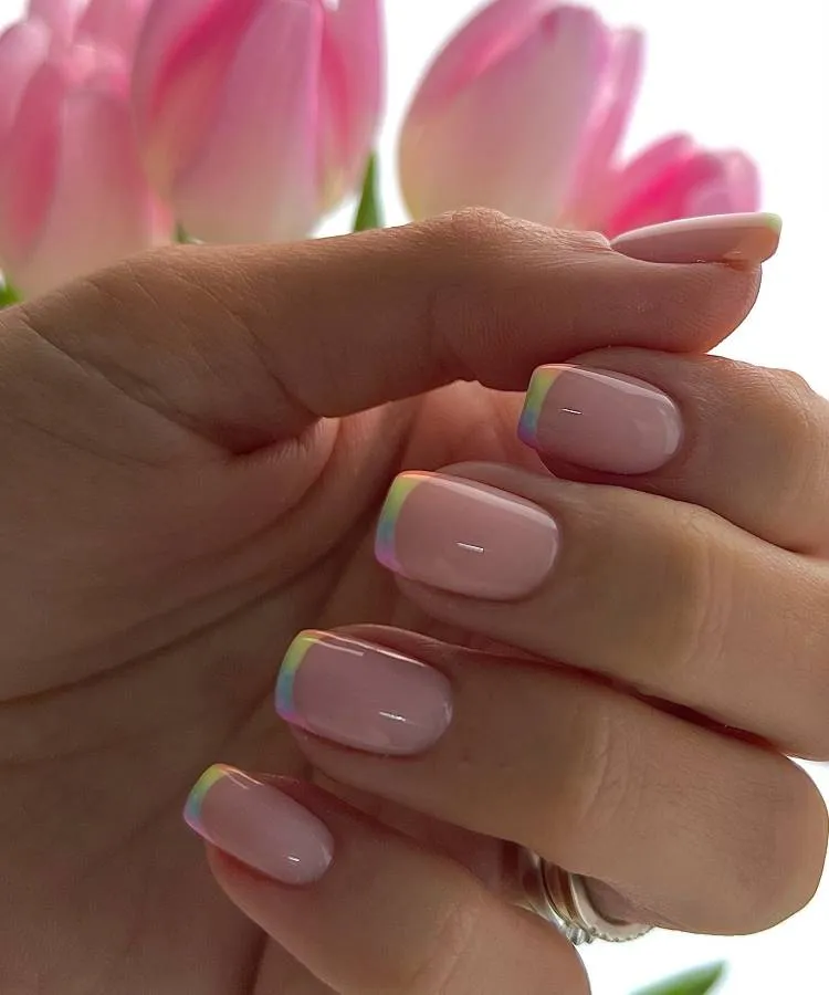 A imagem mostra um close-up de uma mão com unhas bem cuidadas. As unhas estão pintadas com um esmalte rosa claro e apresentam um efeito iridescente sutil que reflete diferentes cores nas pontas, principalmente tons de verde e amarelo. Atrás da mão, há tulipas rosas desfocadas, adicionando um toque estético à imagem.

