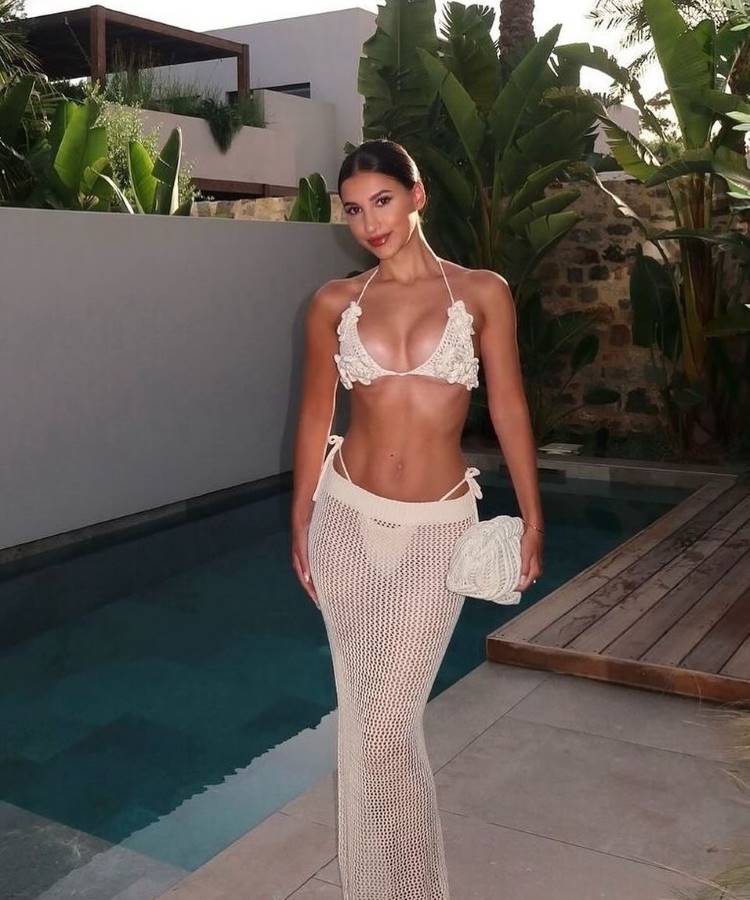Uma mulher com cabelo preso e maquiagem elegante, usa um biquíni e uma saia longa de crochê brancos. Ela posa ao lado de uma piscina, segurando uma pequena bolsa de mão de crochê, em frente a um cenário de vegetação tropical.