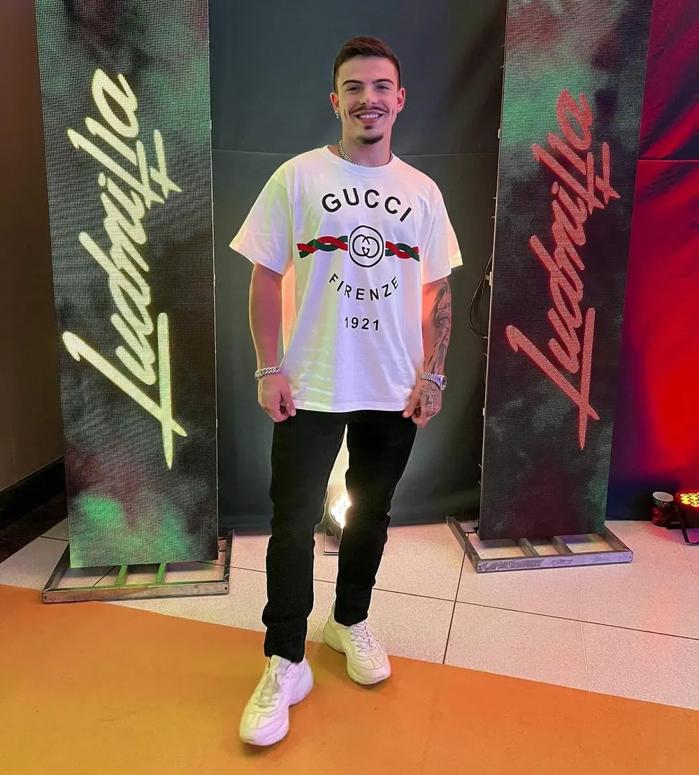 Foto de Thomaz Costa em um evento da Ludimilla. O ator está sorrindo, vestido com uma blusa da Gucci, calça preta e tênis branco.