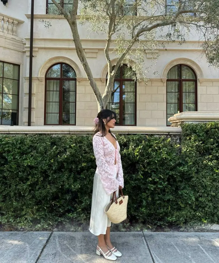 Uma mulher de perfil em frente a um prédio bege com janelas de arcos, ladeado por um arbusto. Ela usa uma blusa de manga comprida rosa de renda, uma saia branca esvoaçante, sapatos de salto brancos e uma bolsa de palha.