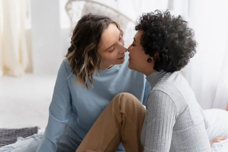 Duas pessoas que se identificam como mulheres, uma de pele clara e cabelos castanhos e outra de pele negra e cabelo preto quase se beijando