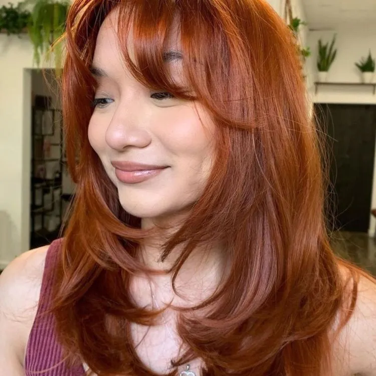 uma mulher ruiva posa virada de lado com uma blusa rosa, seu cabelo médio  é bem repicado e em camadas