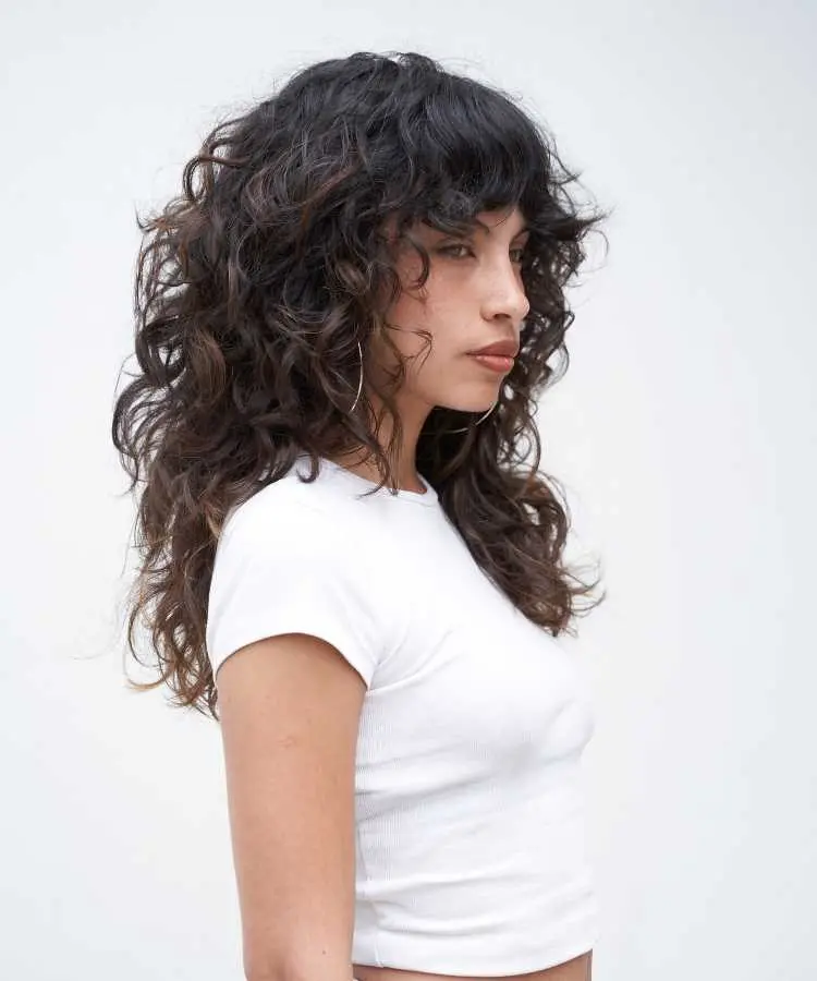 Mulher com cabelos longos, cacheados, com corte shaggy e franja cortininha, usando uma blusa branca ajustada. Ela olha para o lado com expressão serena, destacando o volume e movimento dos fios contra um fundo claro e minimalista.